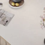 Caesarstone Quartz 4011 Cloudburst Concrete surface with Open magazine, wooden bowl with lemons, and pink flowers in a black vase on white table.