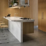 Caesarstone ICON 5131 Calacatta Nuvo surface in Modern kitchen with wooden cabinets, marble island, bar stools, built-in ovens, and a large plant.