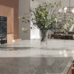 Caesarstone 5133 Symphony Grey surface with A glass vase with leafy branches sits on a marble kitchen island. In the background, a dining table with chairs and modern pendant lights are visible, all in a softly lit kitchen.