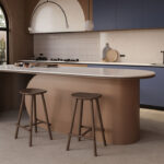 Caesarstone ICON 5144 Rossa Nova surface in Modern kitchen with sleek island, two stools, arched window, blue cabinets, and minimal décor.