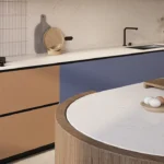 Caesarstone ICON 5144 Rossa Nova surface in Modern kitchen with beige and blue cabinets, countertop stove, sink, round table, and fruit bowls.