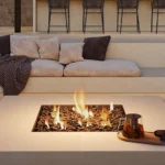 Caesarstone Porcelain 412 Beige Ciment surface in cozy outdoor seating with fire pit, cushioned sofa, and bar stools against stone wall backdrop.